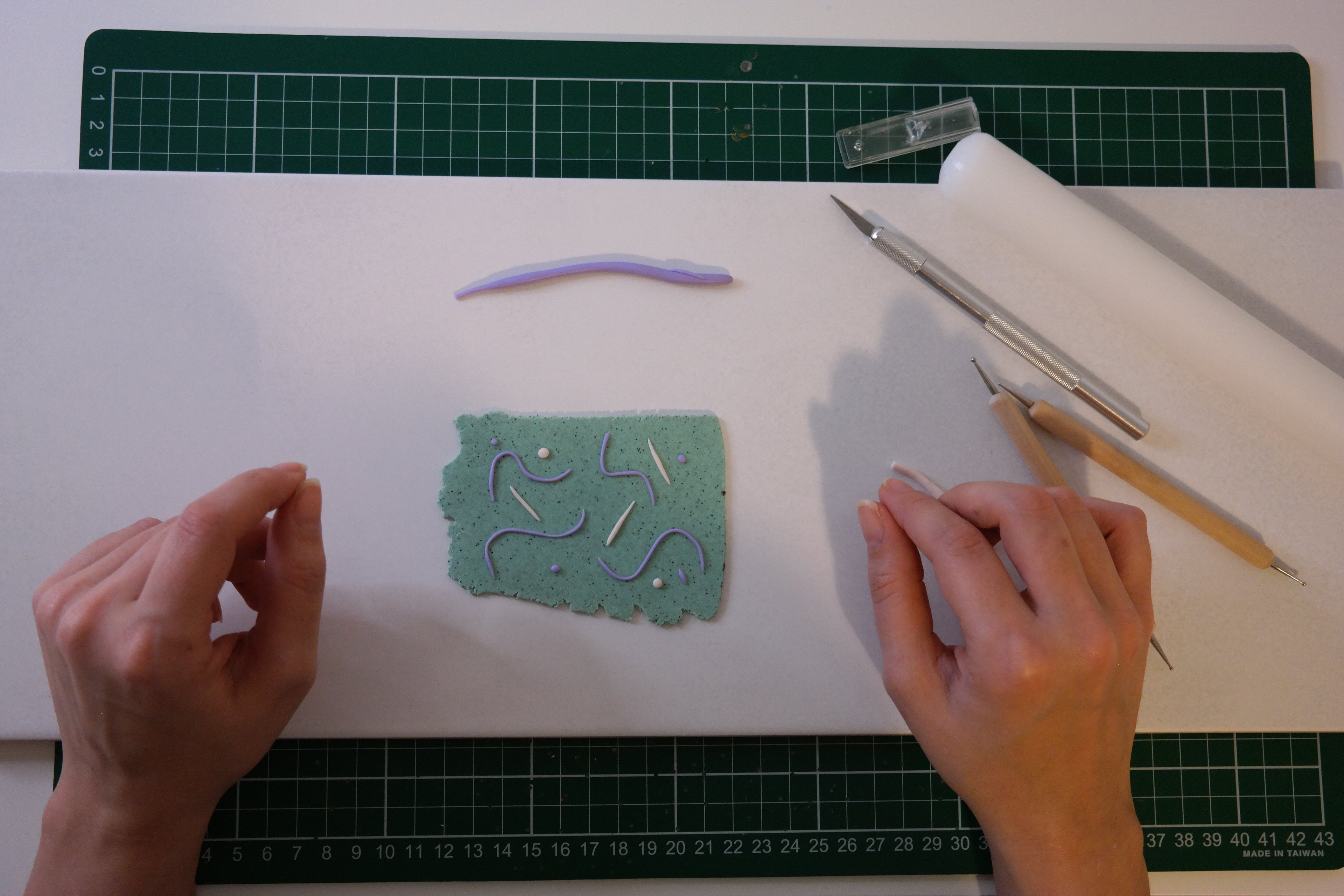 Hands working with clay on a white surface with tools and a green cutting mat.